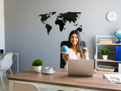 Woman sits at a wooden desk in a modern office, holding a globe and smiling at the camera, with a laptop and a world map on the wall behind her.