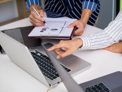 Two professionals review charts on a clipboard while one points at an open laptop.